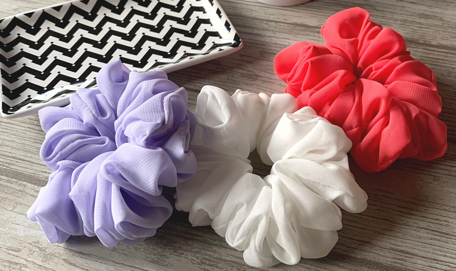 Produktbild med lavendel, vit och korall scrunchie liggandes på bänken med en svart/vit zigzag randigt fat bakom